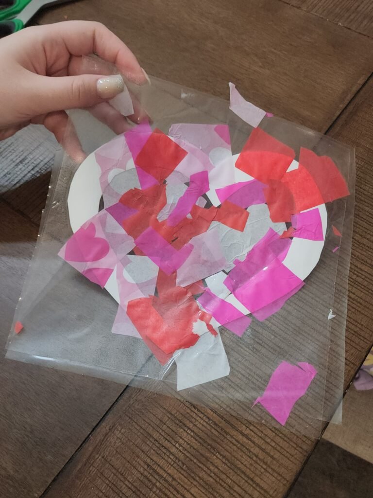 DIY Valentine’s Day craft: Parent and child smoothing out a sheet of contact paper over the tissue paper design