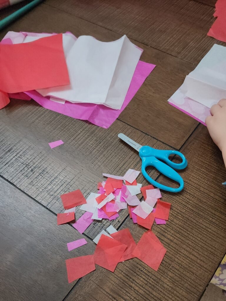 Valentine’s Day craft ideas: Child cutting colorful tissue paper into small squares for a DIY sun catcher project.